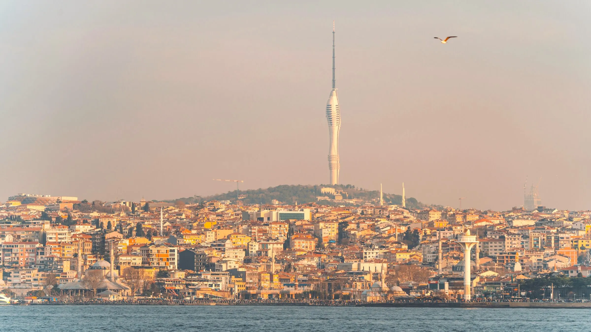 Camlica Tower rising from a green hilltop above the densely built waterfront of Uskudar, seen across the Bosphorus in warm evening light